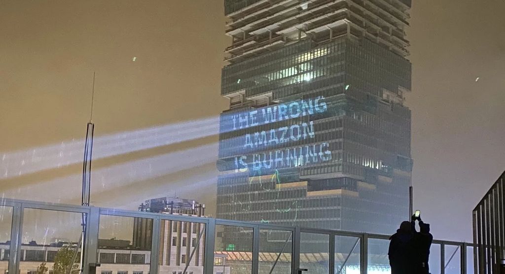 "The Amazon Tower in Berlin by night. A powerful projector projects «The wrong Amazon is burning» on its side."