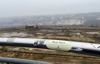 "View into the coal mine Garzweiler. In the front you can see a guard rail on which someone put a sticker reading «Nett hier». The background is blurry and shows the mine itself. Everything is brown, all shades of brown. The sky is cloudy and the view partially obscured by layers of fog."