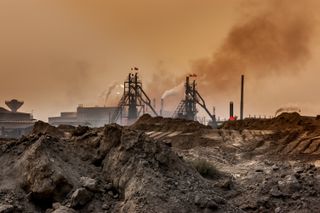 "A mining field, the foreground are heaps of dirst, possibly minerals dug out from the ground. In the center of the image there are two towers, flying the Chinese flag. They are surrounded by other buildings and smoking chimneys. The sky has a dark orange, toxic tint."